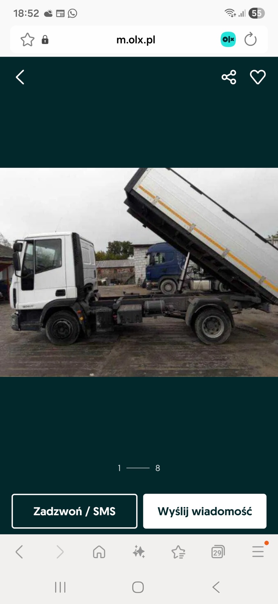 Biała wywrotka z podniesioną skrzynią ładunkową na tle zabudowań. Widoczny fragment kabiny i podwozia. W tle zaparkowany niebieski samochód ciężarowy.