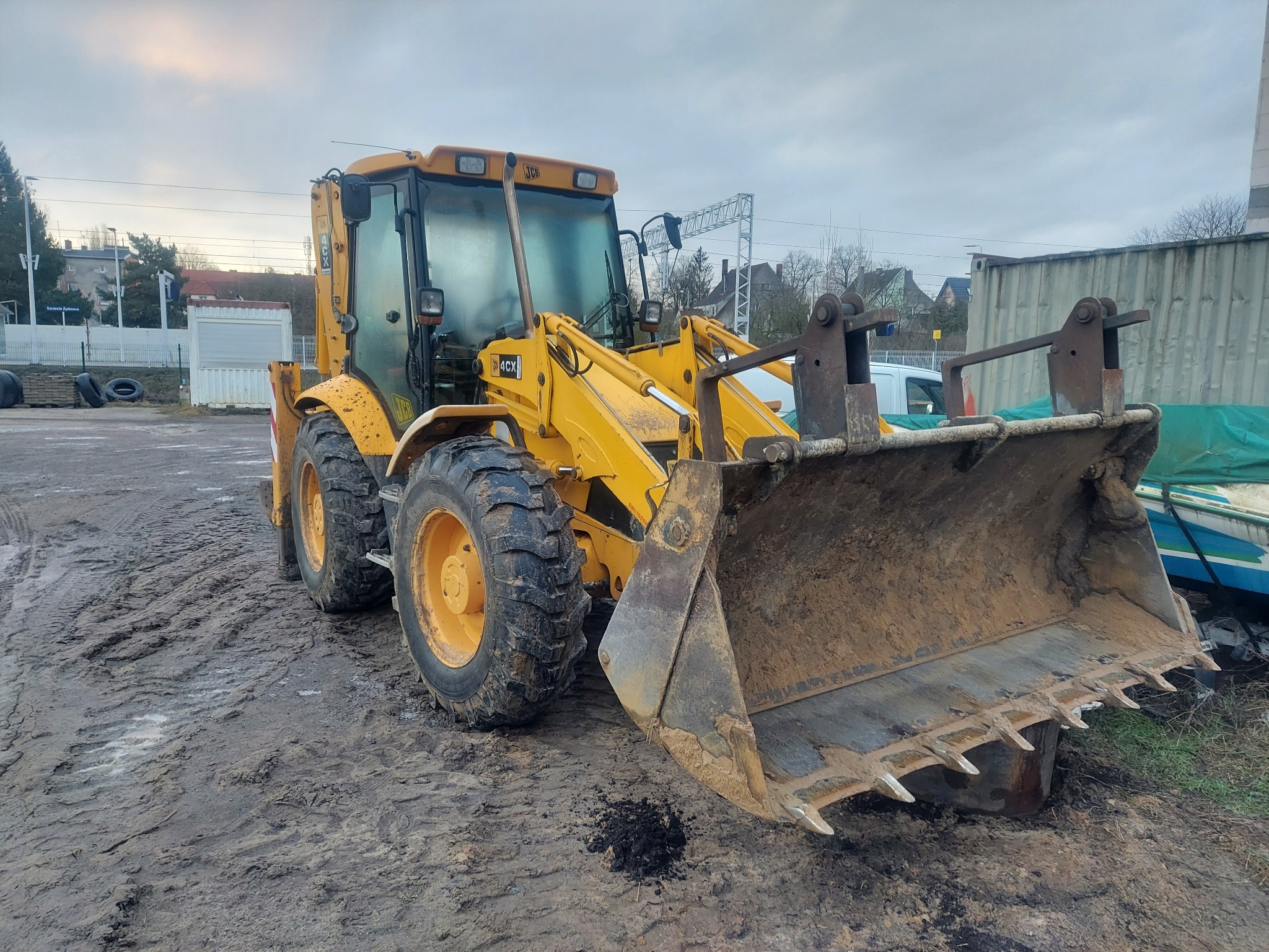 Żółta koparko-ładowarka JCB 4CX z brudną łyżką na błotnistym terenie, w tle budynki i konstrukcja stalowa. Widok z przodu, pochmurne niebo.