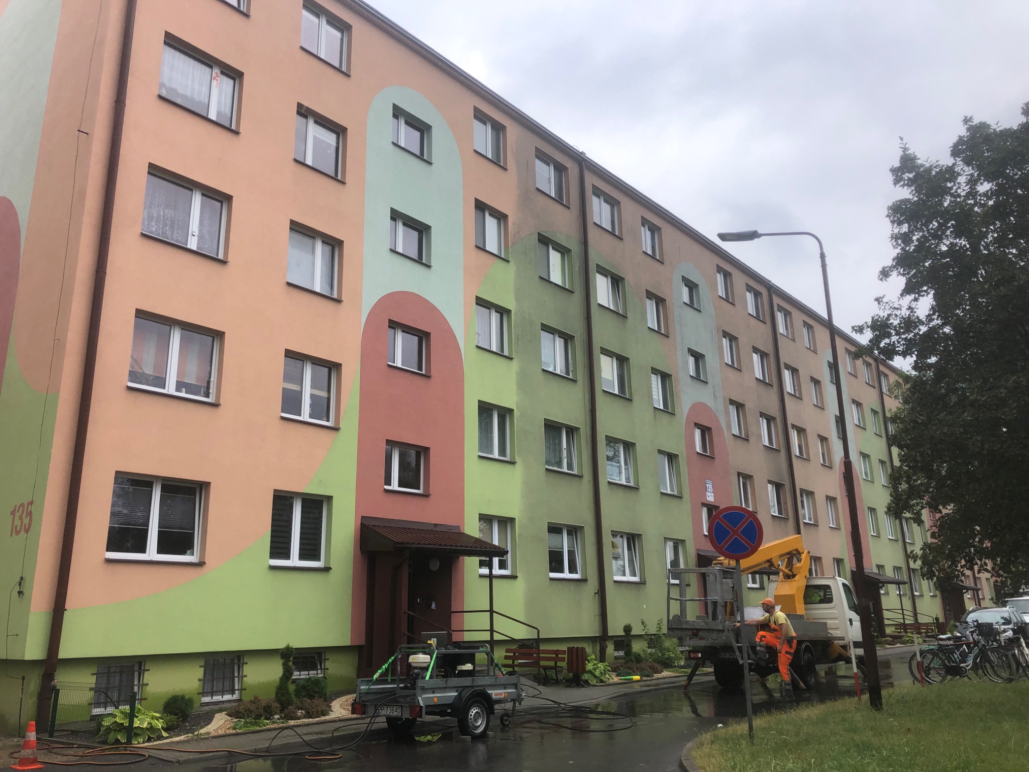 Pracownik w pomarańczowym kombinezonie na podnośniku koszowym, przygotowuje się do pracy przy wielobarwnej elewacji bloku mieszkalnego, widoczny sprzęt czyszczący na przyczepce.