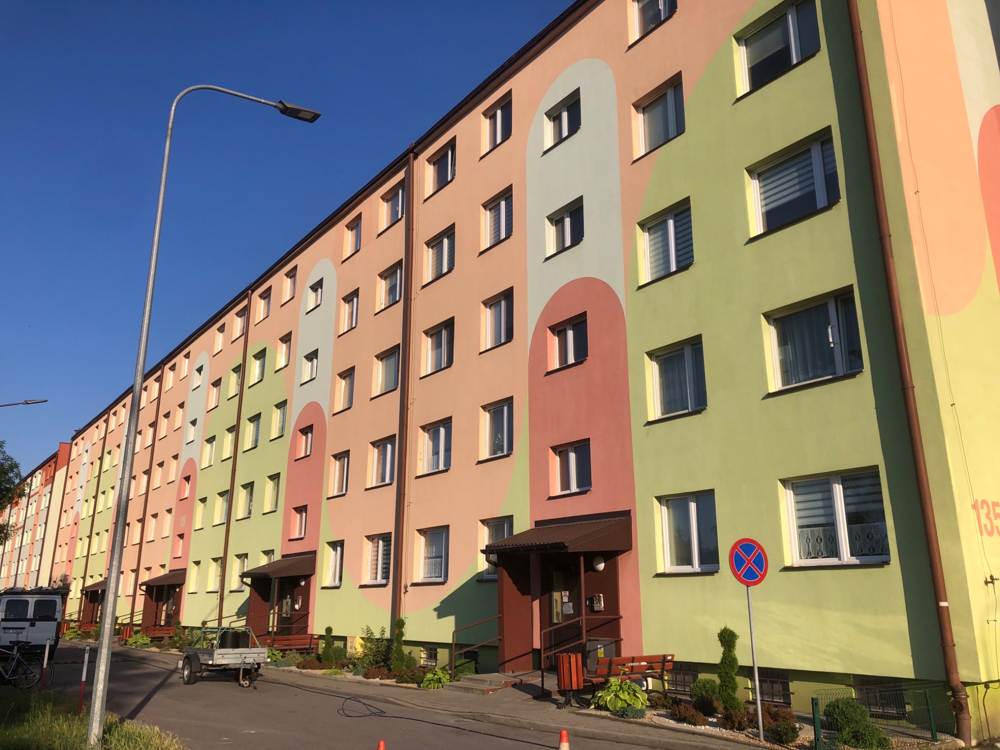 Wielopiętrowy budynek mieszkalny z odnowioną, kolorową elewacją w odcieniach zieleni, różu i beżu, widziany pod kątem z ulicy z zaparkowanym samochodem dostawczym i przyczepką.