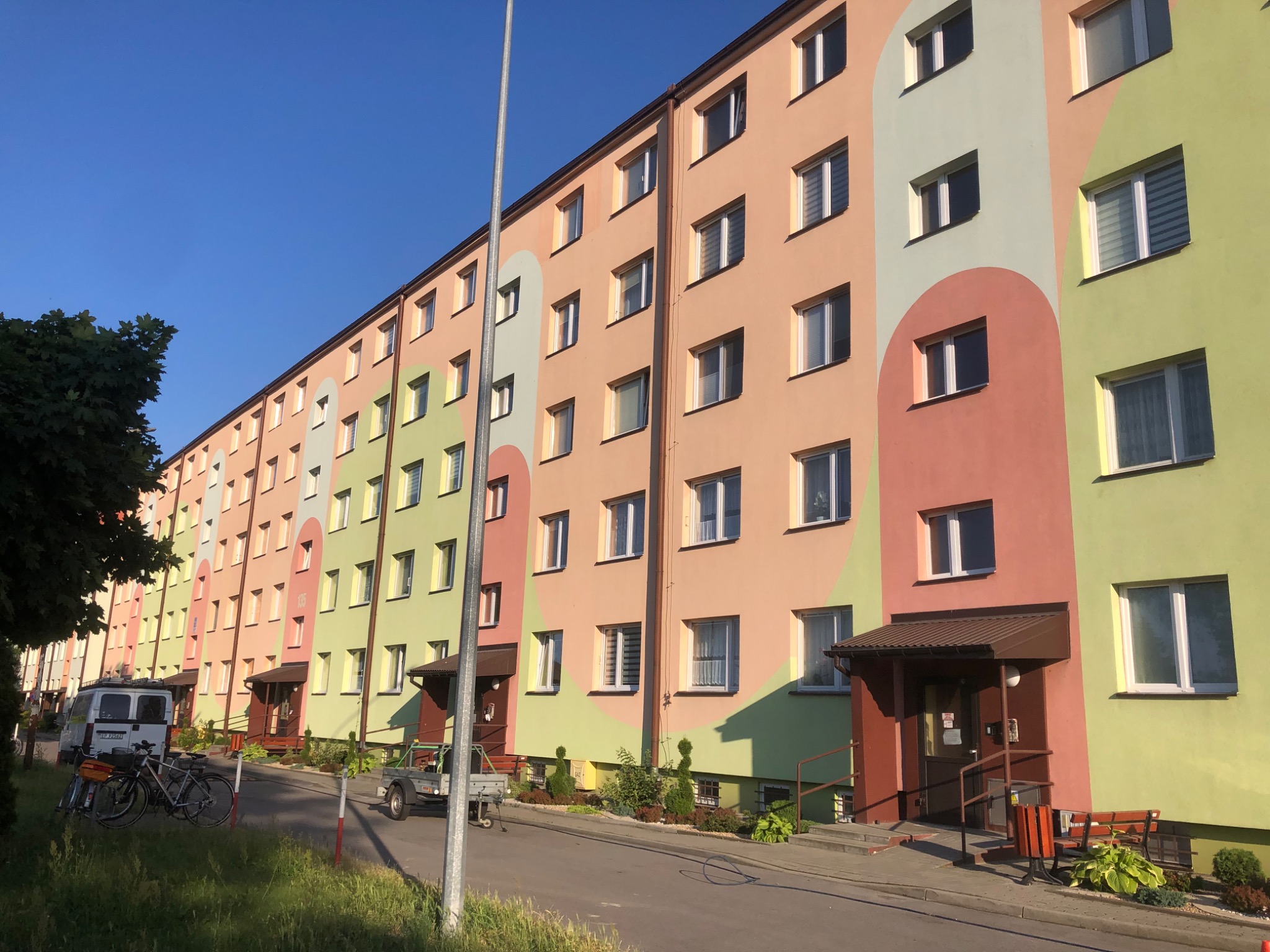 Wielopiętrowy budynek mieszkalny z odnowioną elewacją w pastelowych kolorach (różowy, zielony, beżowy) z geometrycznymi zdobieniami w kształcie półokręgów, widok z poziomu ulicy w słoneczny dzień.