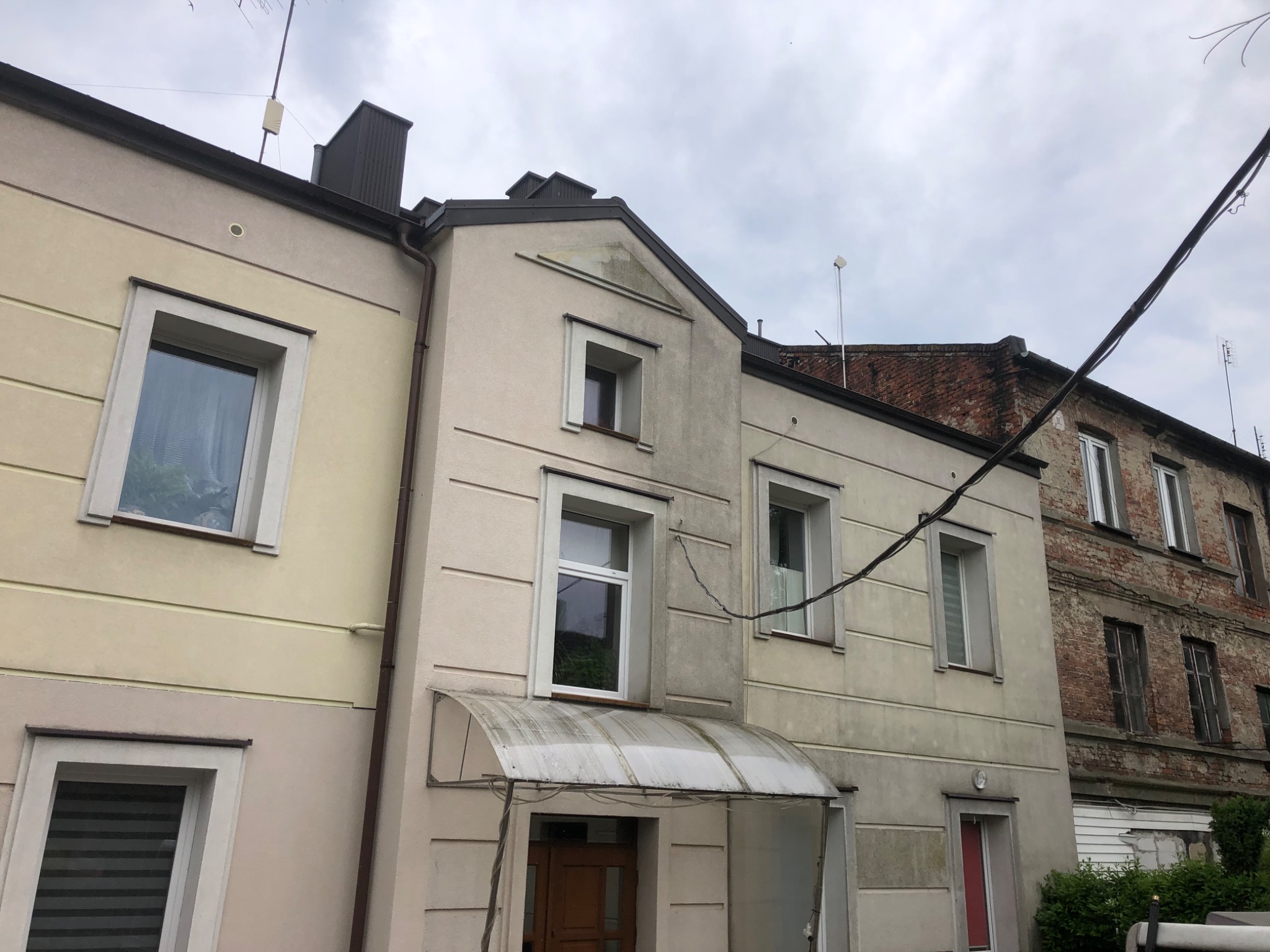 Dwupiętrowa kamienica z elewacją w odcieniach beżu i szarości, widoczne ślady zabrudzeń na ścianach, plastikowy daszek nad wejściem, sąsiedni budynek z czerwonej cegły.