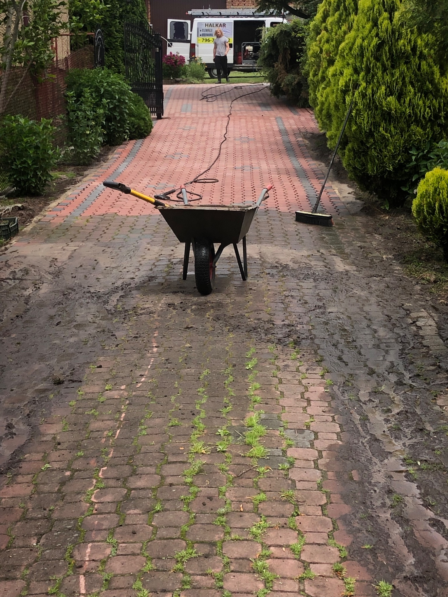 Częściowo umyty chodnik z kostki brukowej, taczka z narzędziami na pierwszym planie, w tle bus firmy sprzątającej i osoba z wężem ciśnieniowym.