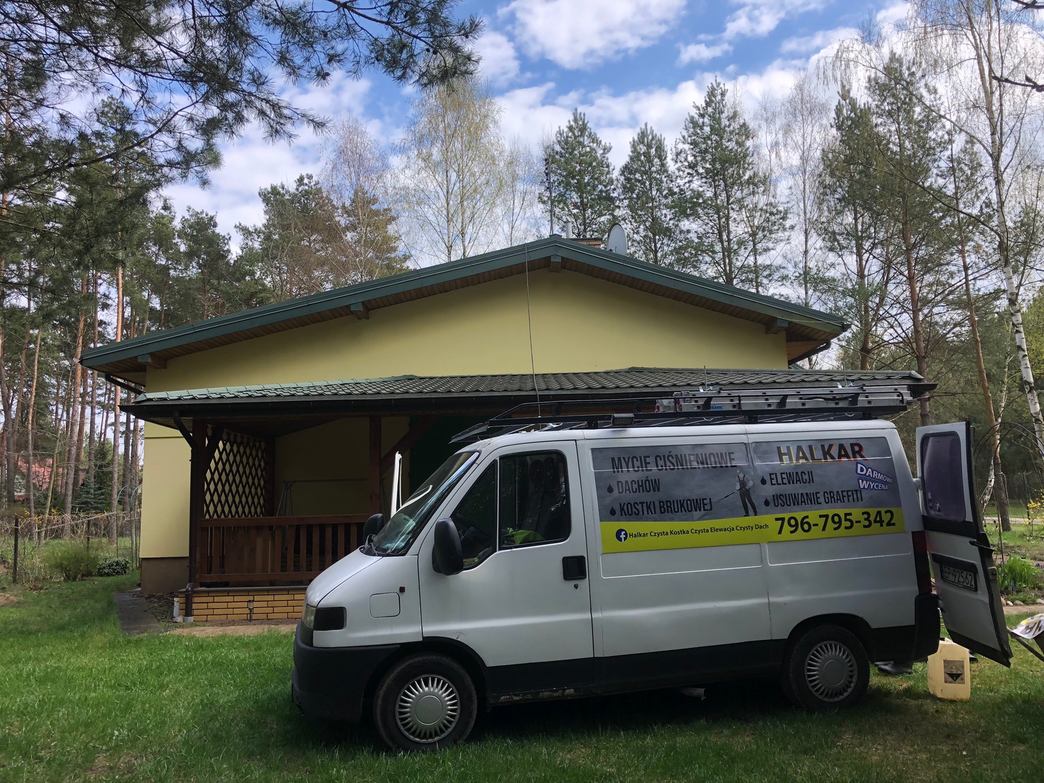 Żółty dom z zielonym dachem widziany zza białego samochodu dostawczego firmy świadczącej usługi czyszczenia ciśnieniowego, z widocznym numerem telefonu i hasłami reklamowymi na karoserii.