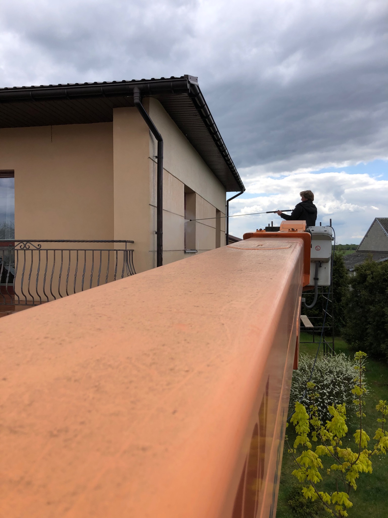 Pracownik z Piotrków Trybunalski myje elewację domu z podnośnika koszowego za pomocą myjki ciśnieniowej. Widok z góry na ramię podnośnika, w tle beżowa elewacja i pochmurne niebo.