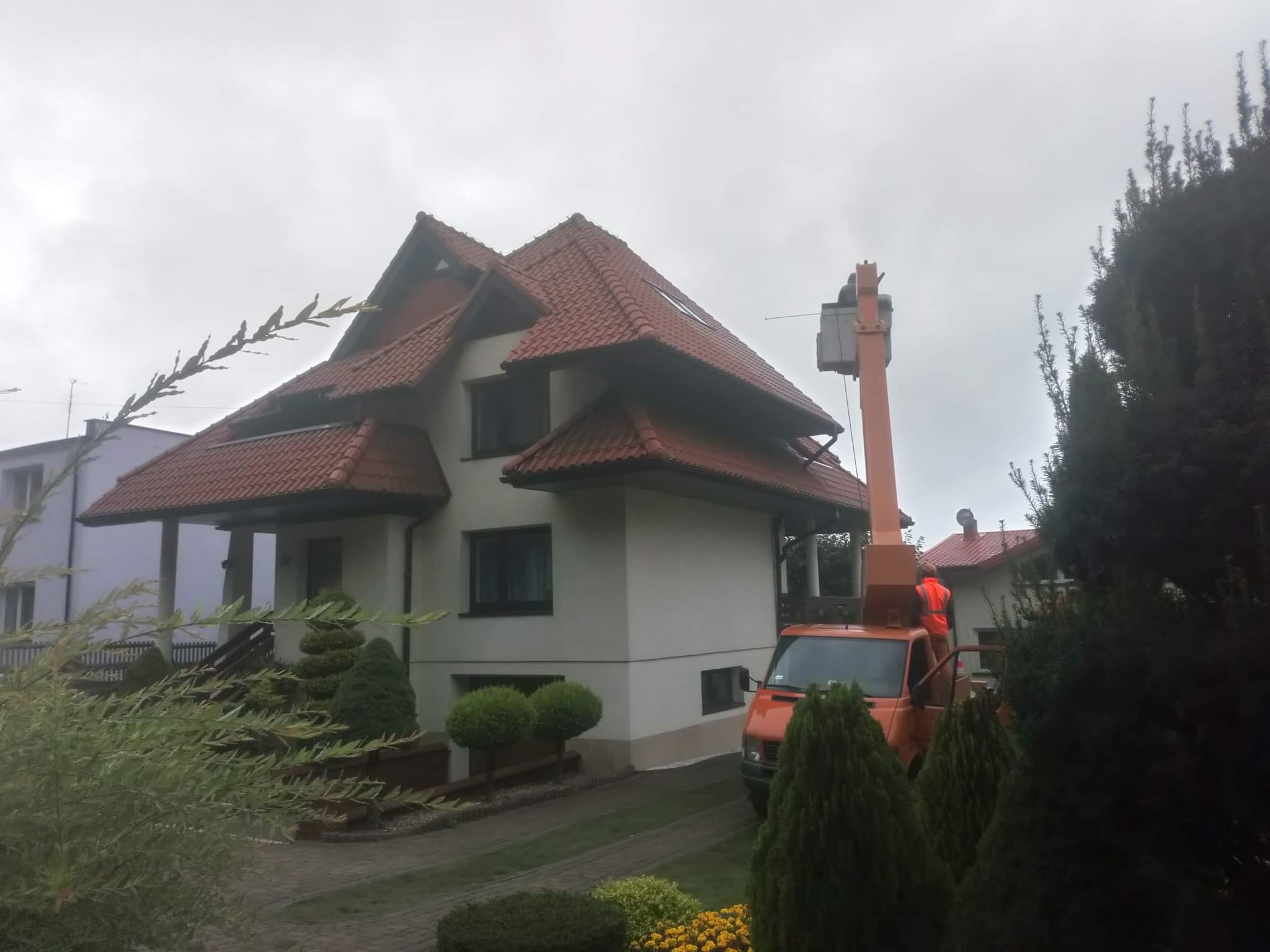 Pracownik w pomarańczowej kamizelce na podnośniku koszowym przy dwupiętrowym domu z czerwoną dachówką, pochmurne niebo w tle.