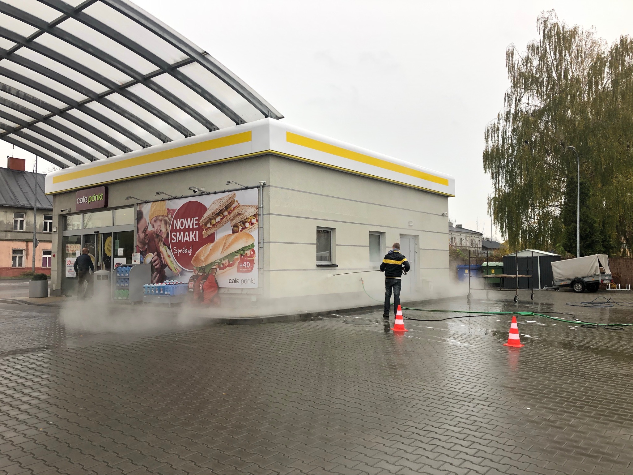 Pracownik w czarnej kurtce z żółtymi paskami myje elewację budynku Cafe Punkt za pomocą myjki ciśnieniowej, widoczne dwa pachołki ostrzegawcze i ulotka reklamowa na ścianie.