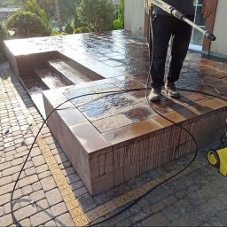 SELBRUK - Czyszczenie ciśnieniowe tarasu z kostki brukowej, widoczny pracownik z lancą myjki Karcher oraz fragment zadbanego ogrodu z tujami i kwiatami.