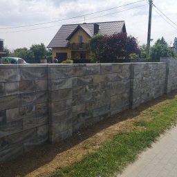 SELBRUK - Nowoczesny, wysoki mur z szarej kostki łupanej, oddzielający posesję z żółtym domem z brązowym dachem, widoczny z chodnika wyłożonego kostką brukową.