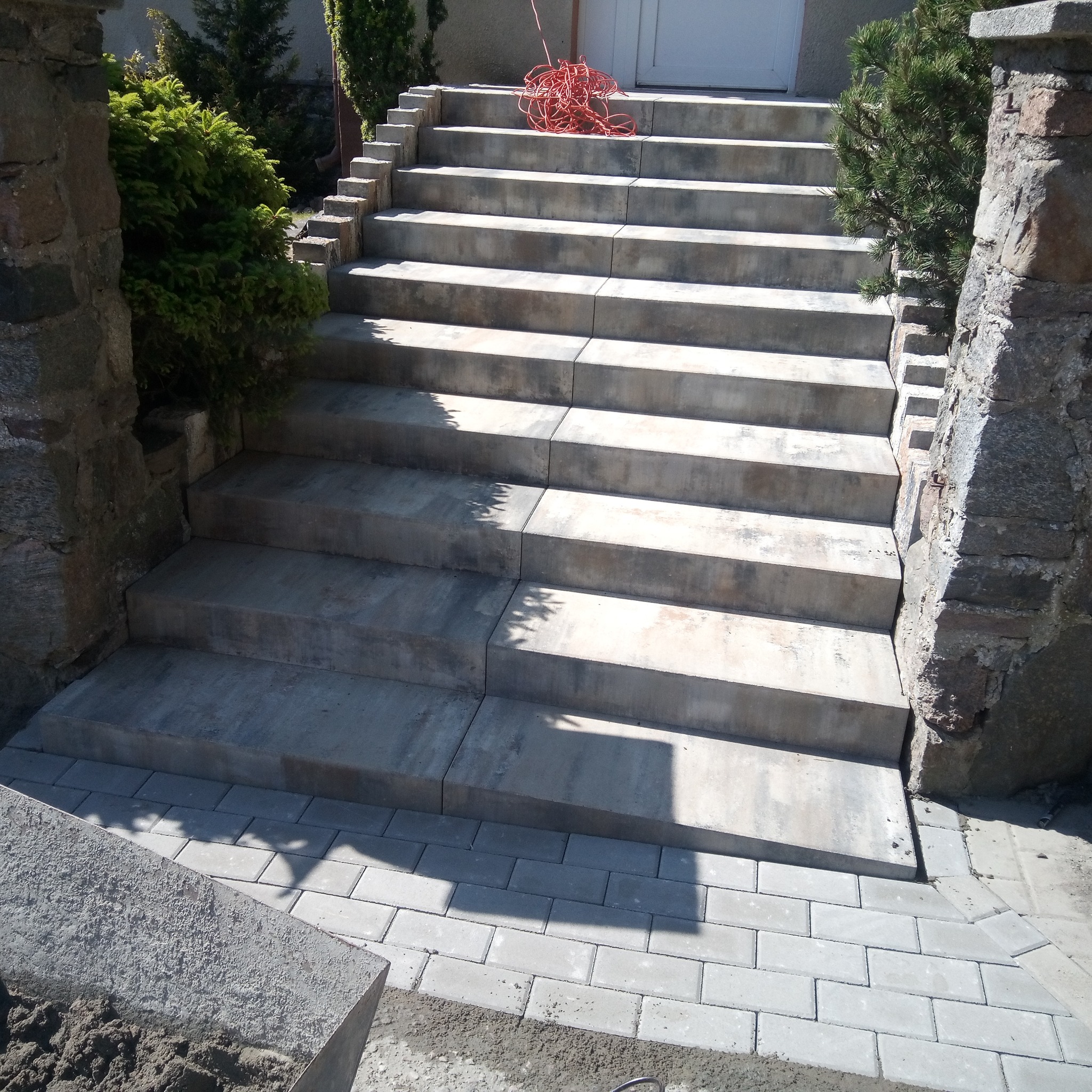 Schody wejściowe wyłożone szarą kostką brukową, obramowane kamiennymi słupkami i zielenią, z pomarańczowym kablem na górnym stopniu.