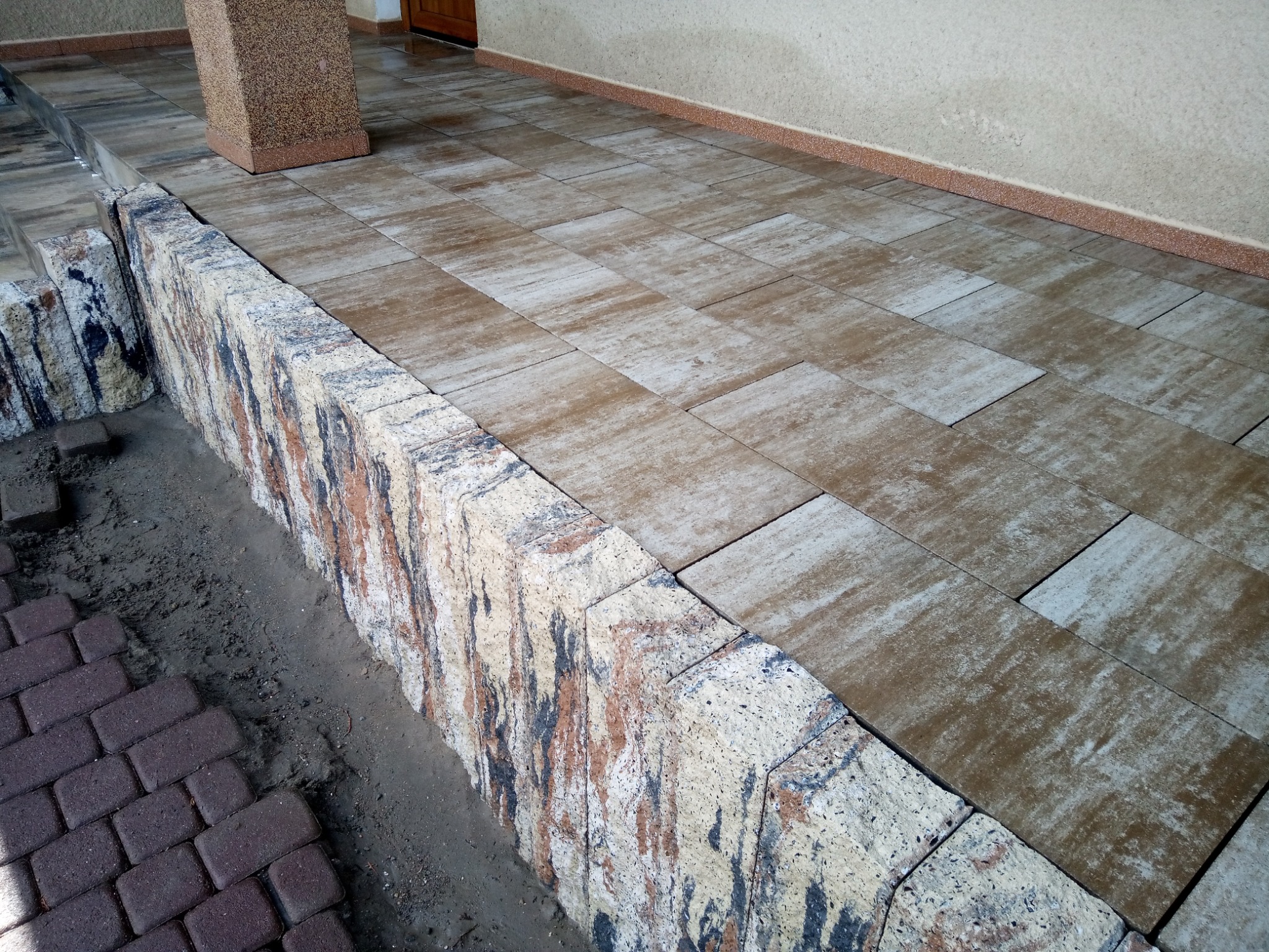 Ułożona nawierzchnia z prostokątnej kostki brukowej w odcieniach beżu i brązu, obrzeże z dekoracyjnej kostki o nieregularnym wzorze, widoczne fragmenty niedokończonej ścieżki z ciemnobrązowej kostki.