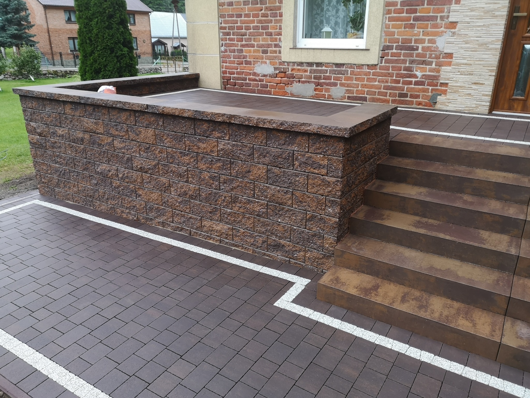 Brązowa kostka brukowa ułożona na podjeździe z białym obramowaniem, schody z kostki prowadzące do drzwi wejściowych, mur z kostki brukowej po lewej stronie.