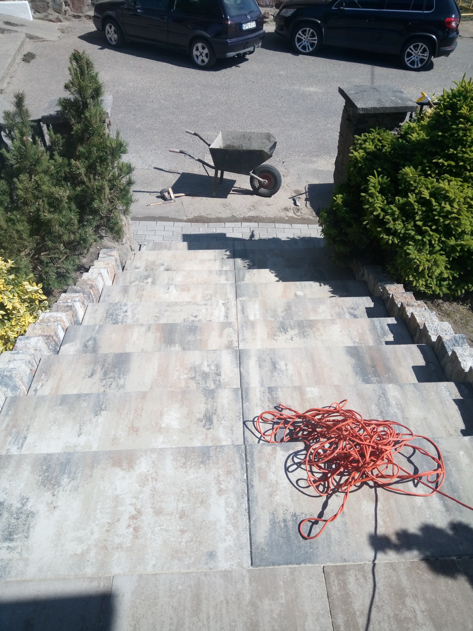 Schody zewnętrzne wyłożone kostką brukową w odcieniach szarości i beżu, widok z góry, na schodach zwinięty pomarańczowy przedłużacz, w tle taczka z zaprawą, samochody i zieleń.