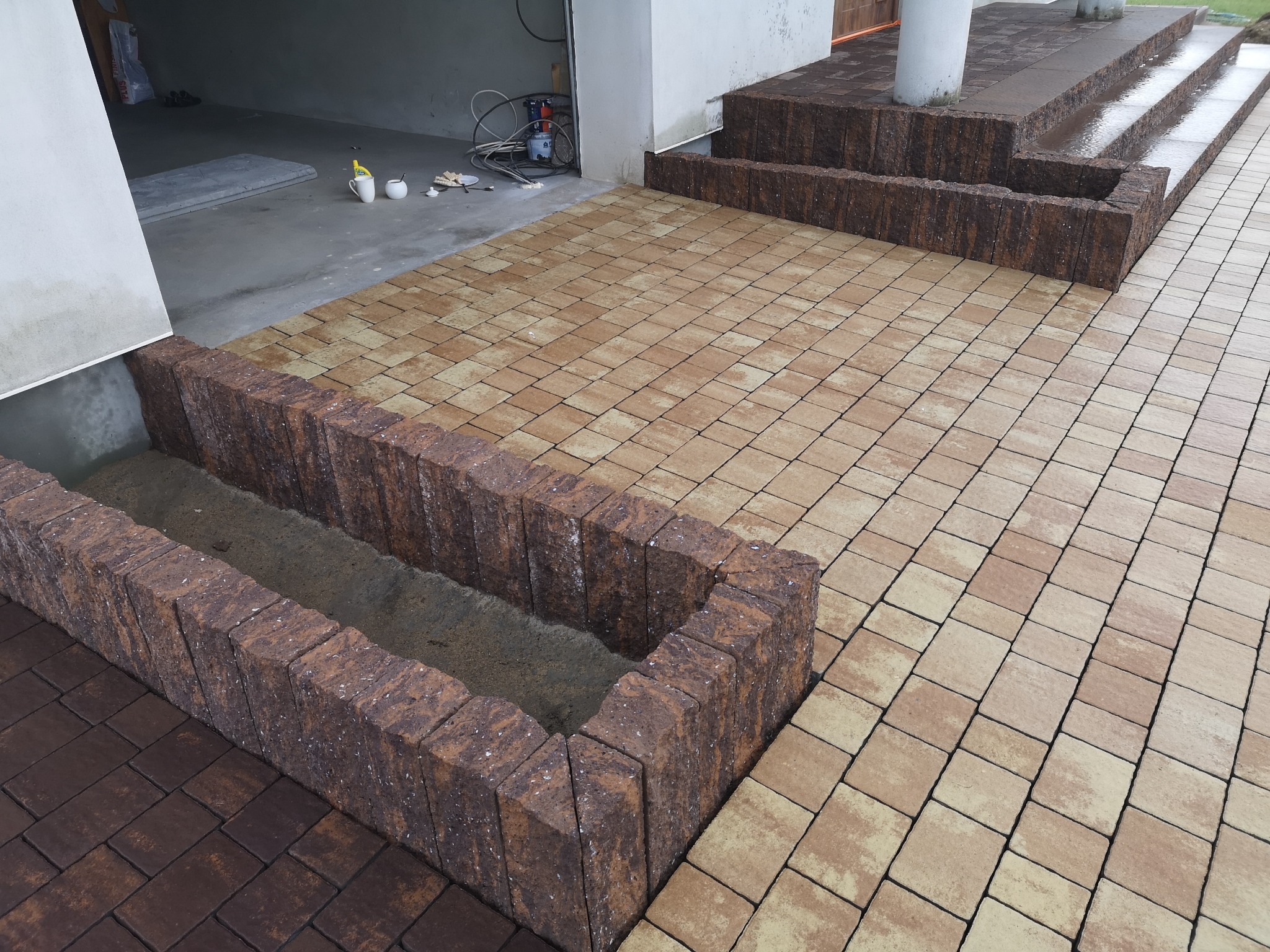 Ułożona kostka brukowa w dwóch odcieniach, beżowym i brązowym, z obrzeżami z kostki w kolorze brązowym, tworzącymi schody i rabatę wypełnioną ziemią, widok z góry.