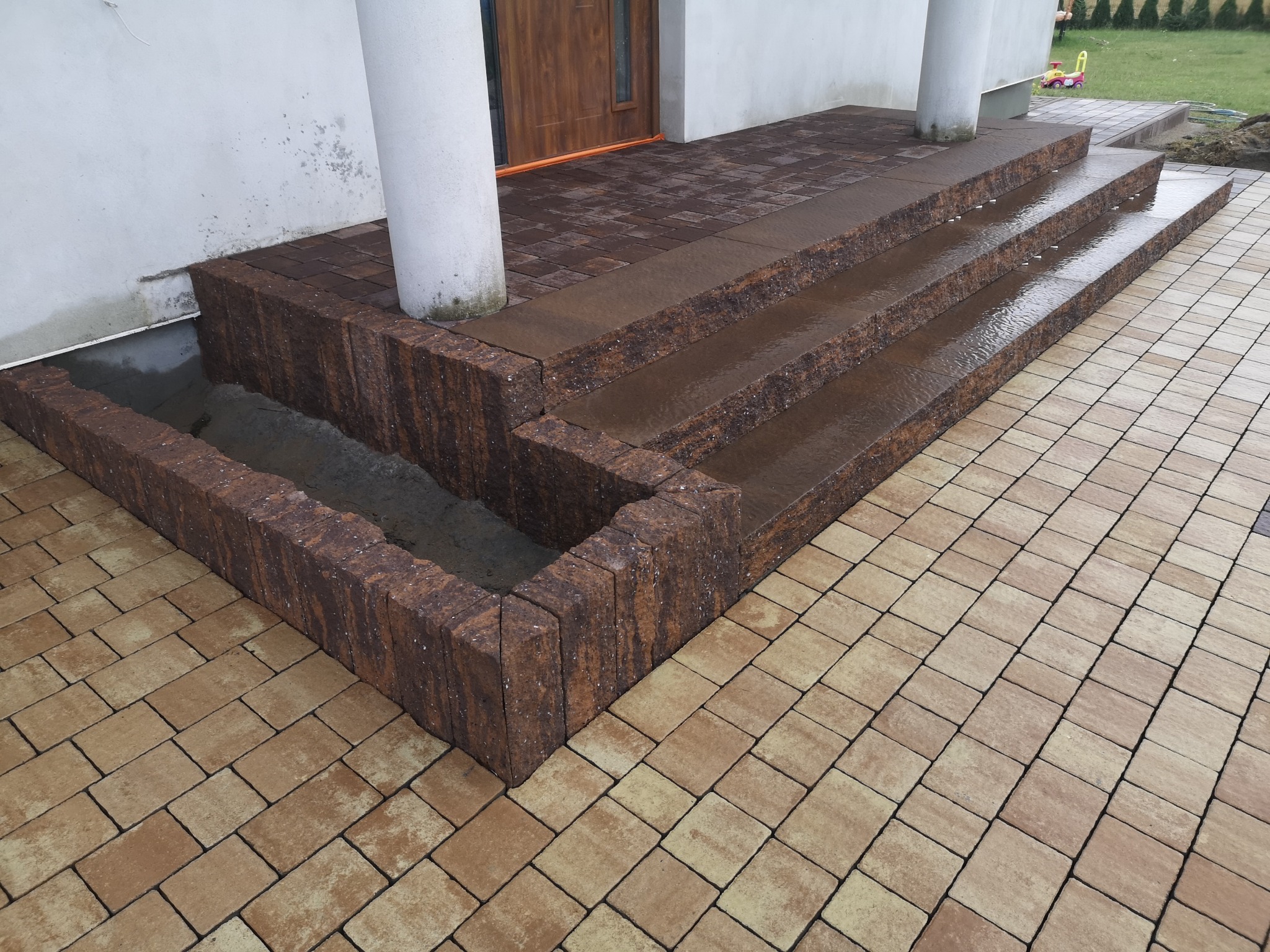 Schody wejściowe do budynku wyłożone kostką brukową w odcieniach brązu i beżu, z widocznymi dwoma kolumnami podtrzymującymi zadaszenie i fragmentem trawnika w tle.
