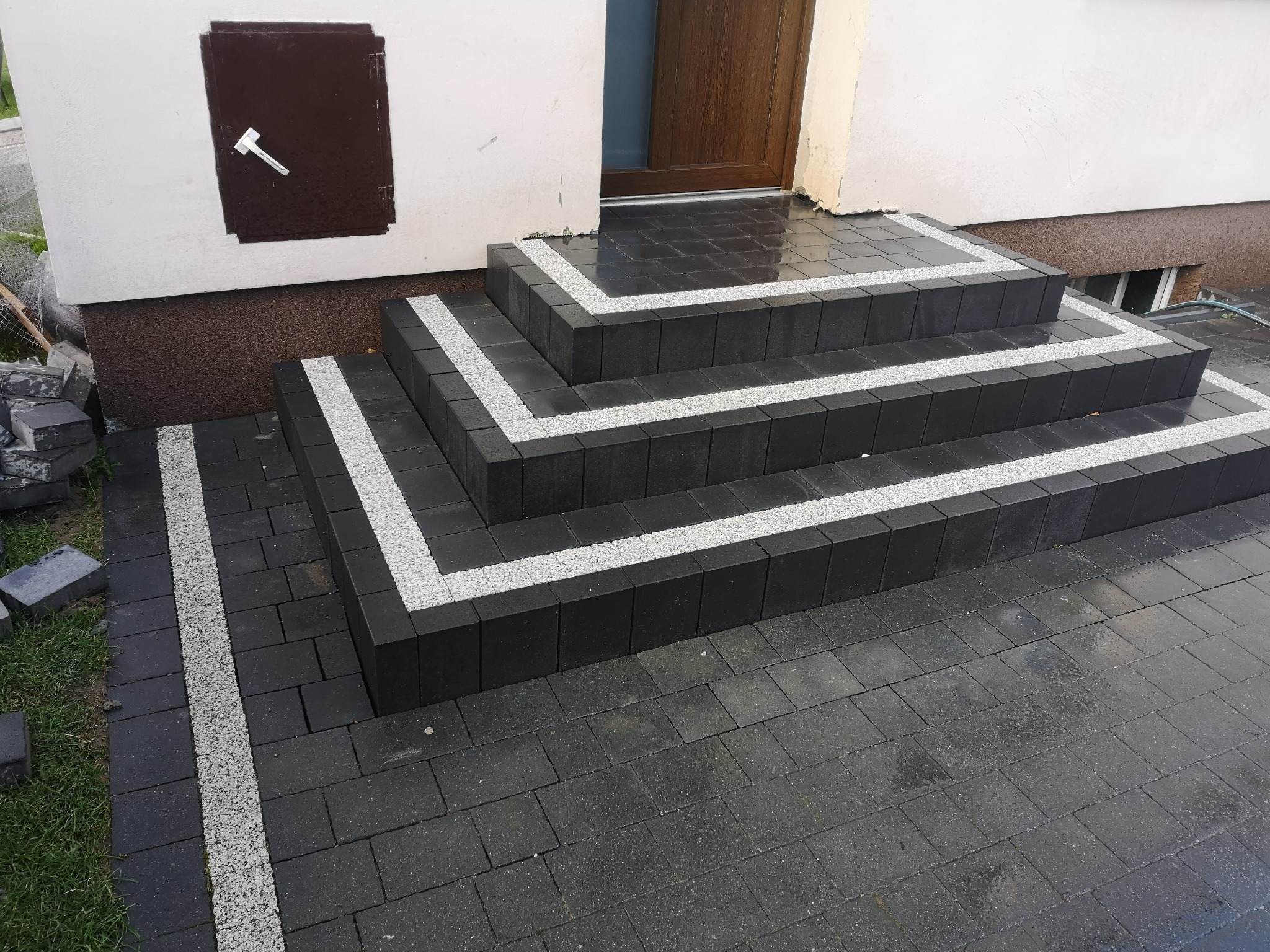 Schody wejściowe wyłożone ciemną kostką brukową z jasnymi, kontrastującymi paskami z kruszywa, prowadzące do drzwi wejściowych budynku z brązową elewacją i małym oknem piwnicznym.