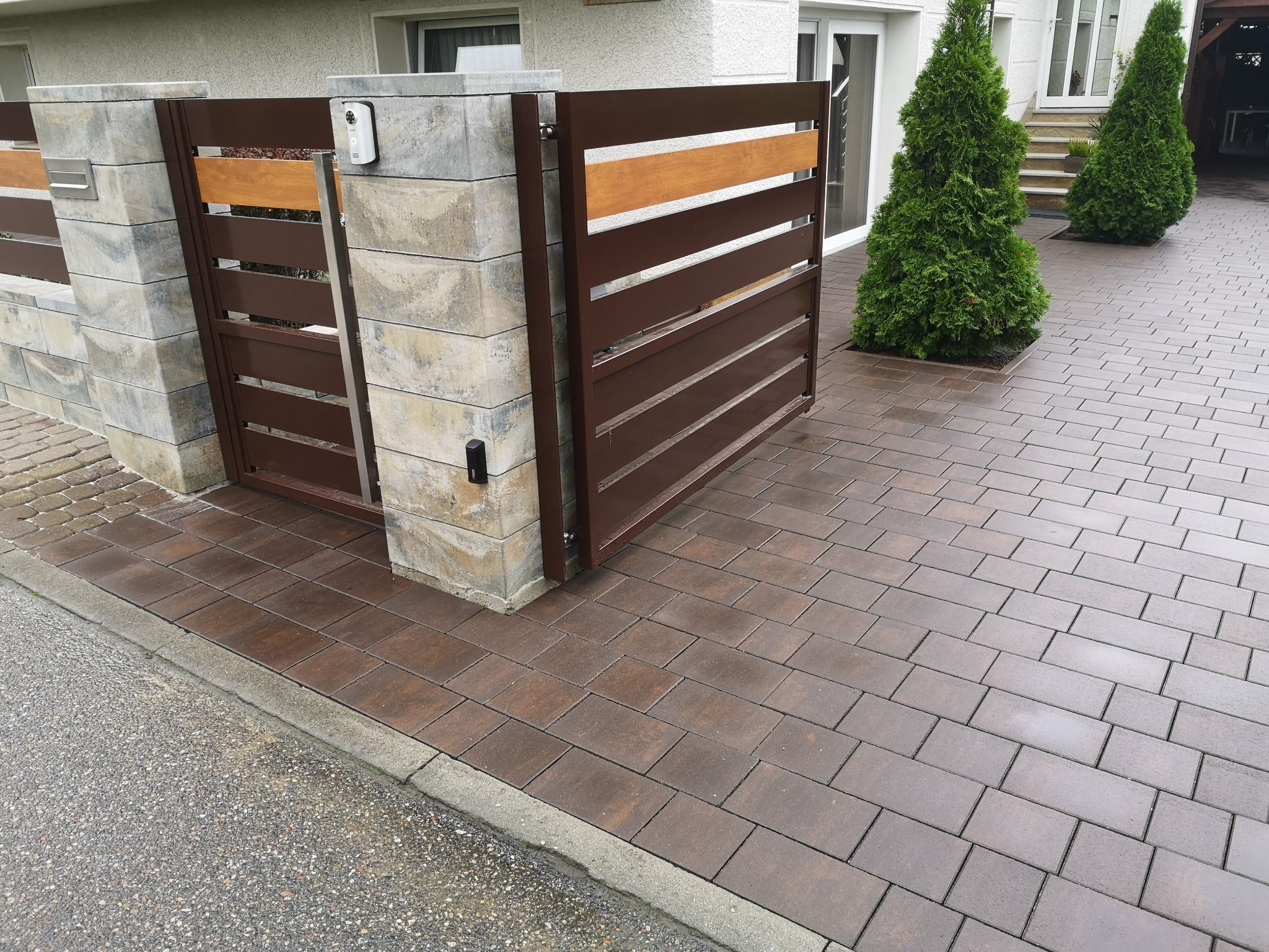 Nowoczesna brama wjazdowa z brązowymi, poziomymi panelami i kamiennymi słupkami, z wideodomofonem, prowadząca na posesję z kostką brukową w odcieniach brązu i dwoma stożkowymi tujami przed wejściem...