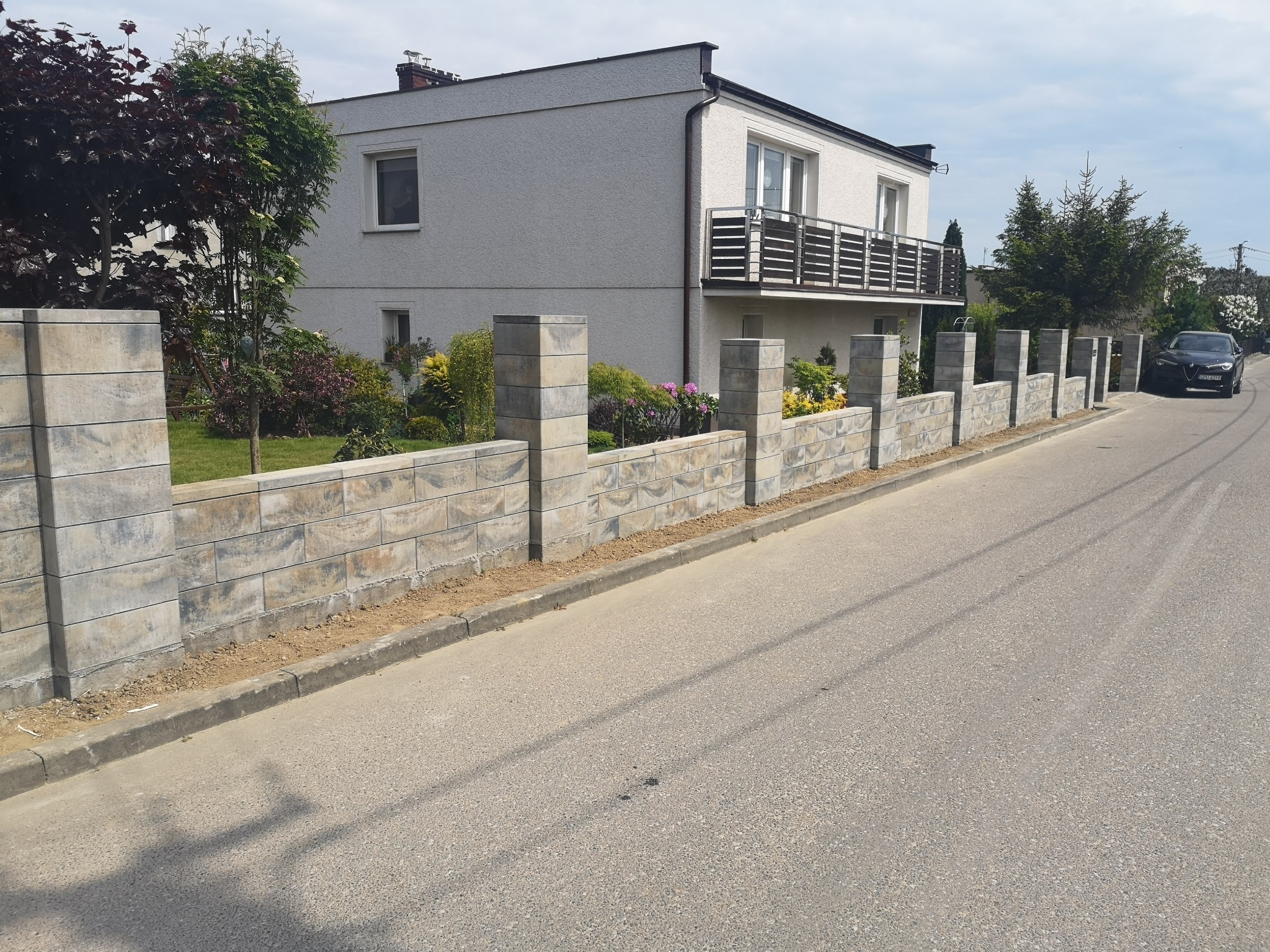 Nowoczesne ogrodzenie modułowe z szarej kostki w Pucku, widok z ulicy, dom z balkonem w tle.