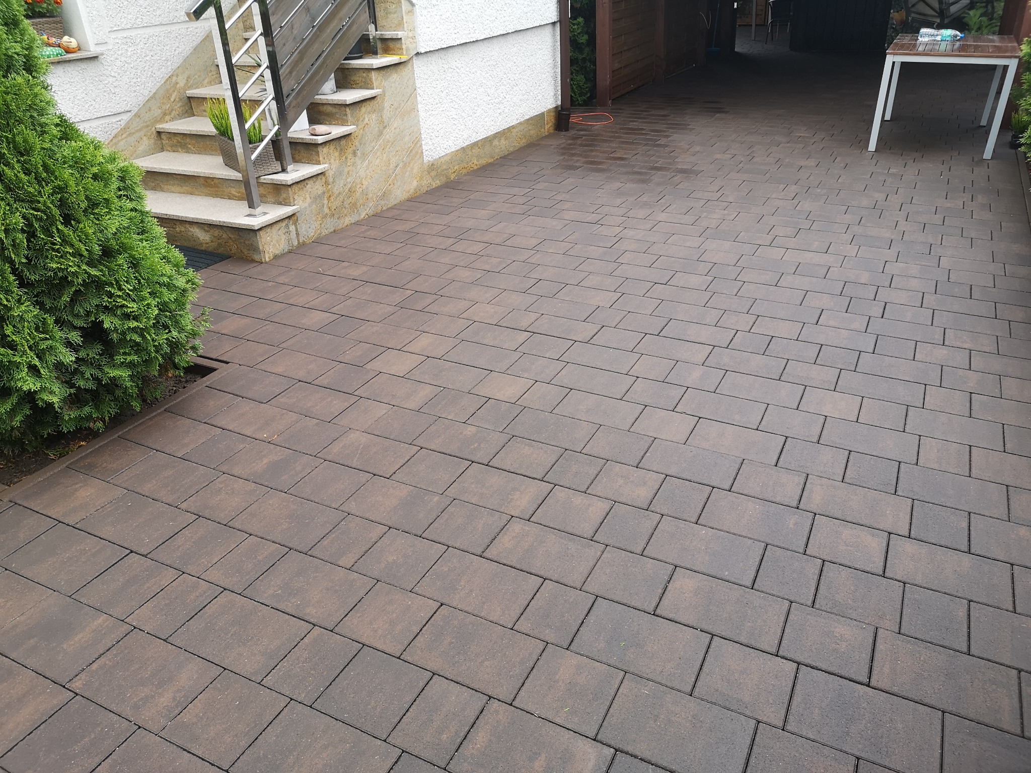 Ułożona, brązowa kostka brukowa na podjeździe, widoczne schody wejściowe do budynku i zielony krzew po lewej stronie. Na kostce widać delikatne ślady wilgoci.