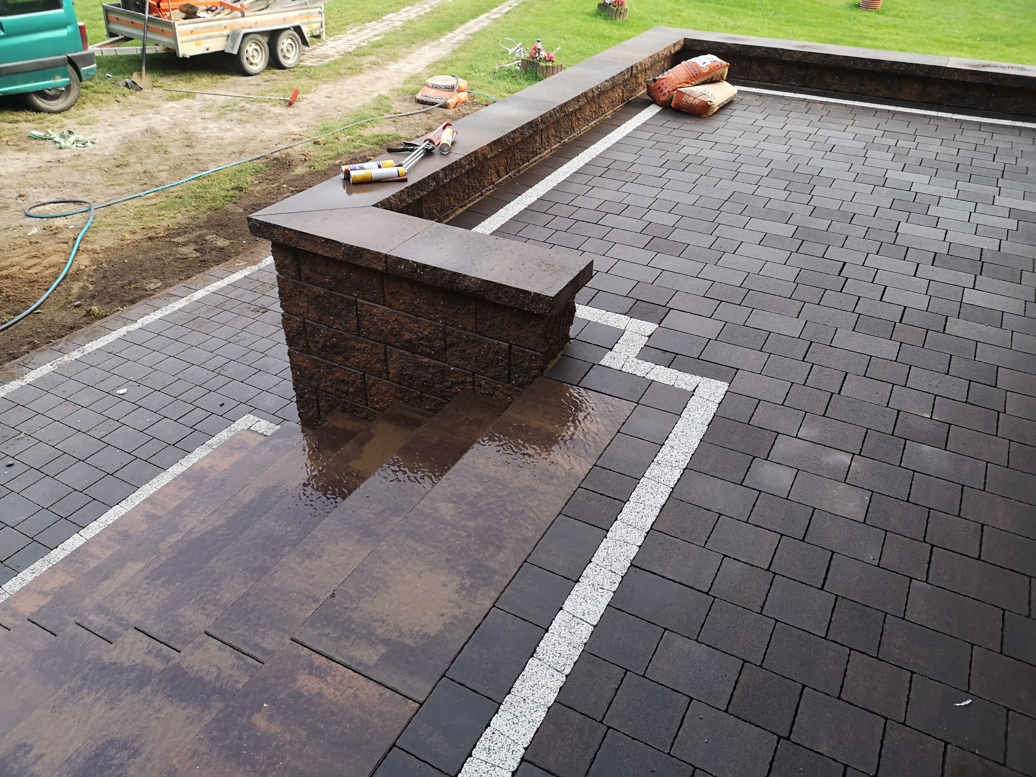 Ułożona, ciemna kostka brukowa z jasnymi fugami i schodami, widoczne narzędzia i worki z materiałami budowlanymi na murku w tle, prawdopodobnie po deszczu.