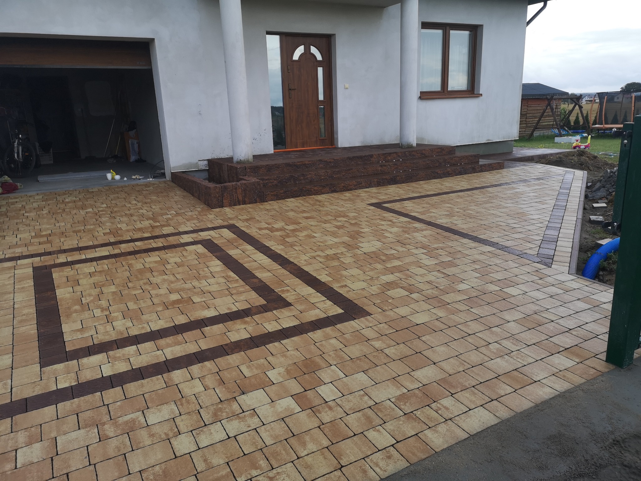 Ułożona kostka brukowa w dwóch odcieniach tworząca dekoracyjny wzór przed domem z garażem; widoczne schody wejściowe również wykończone kostką.