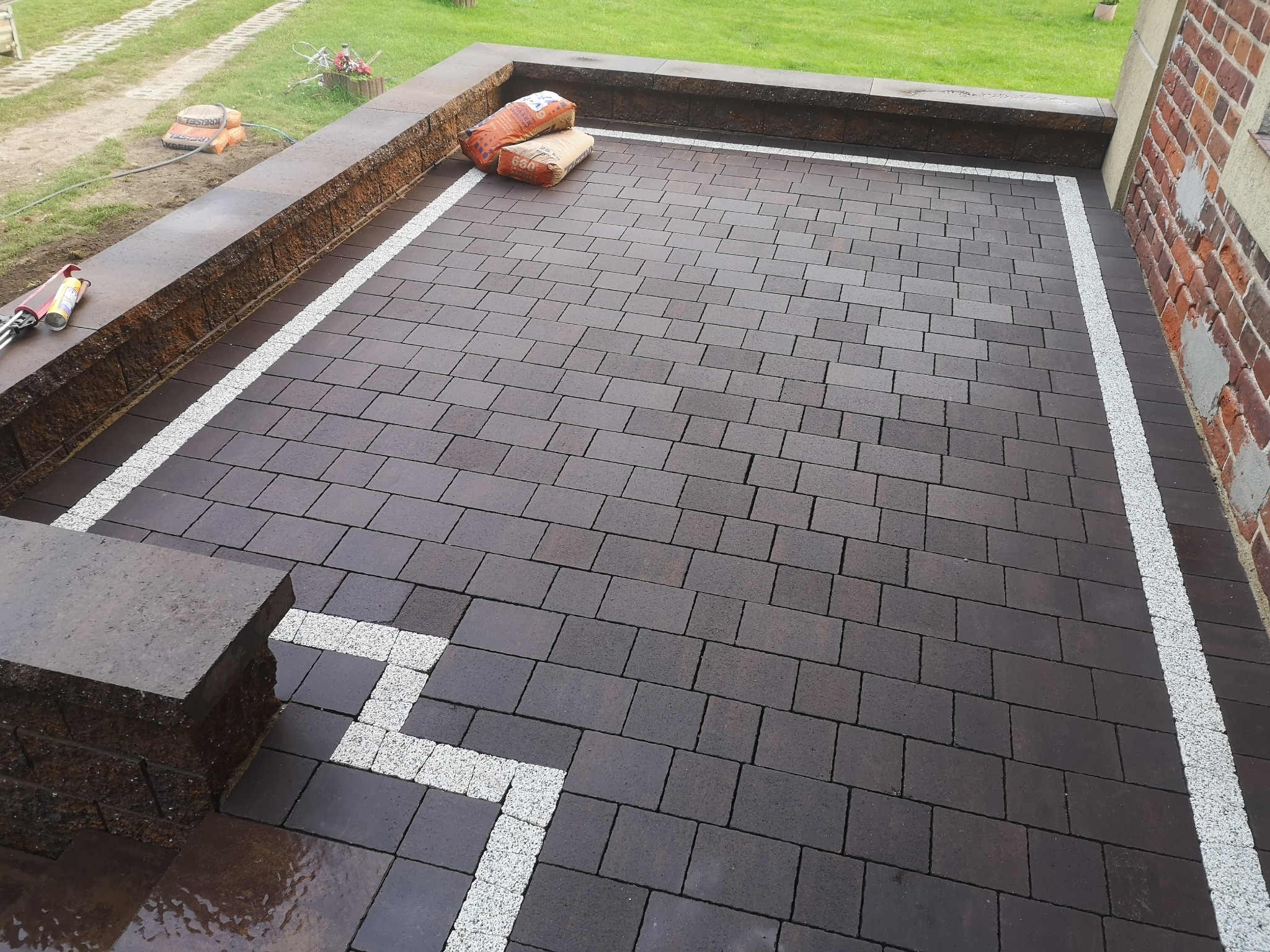 Nowa, brązowa kostka brukowa ułożona na tarasie z dekoracyjnym, białym obramowaniem z grysu; w tle widoczne worki z materiałem budowlanym i fragment budynku z cegły.