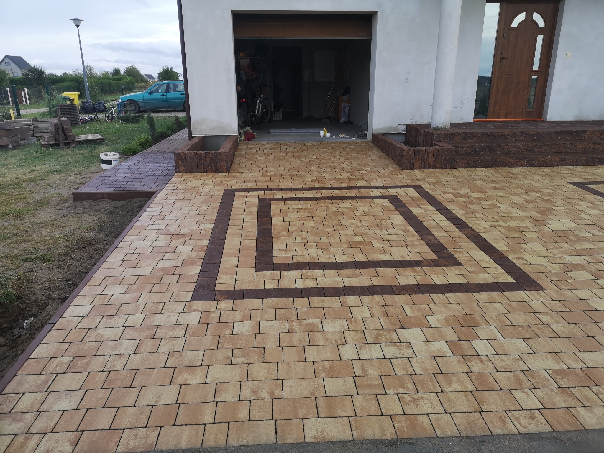 Beżowo-brązowa kostka brukowa ułożona w geometryczny wzór przed domem z otwartym garażem i brązowymi schodami wejściowymi.