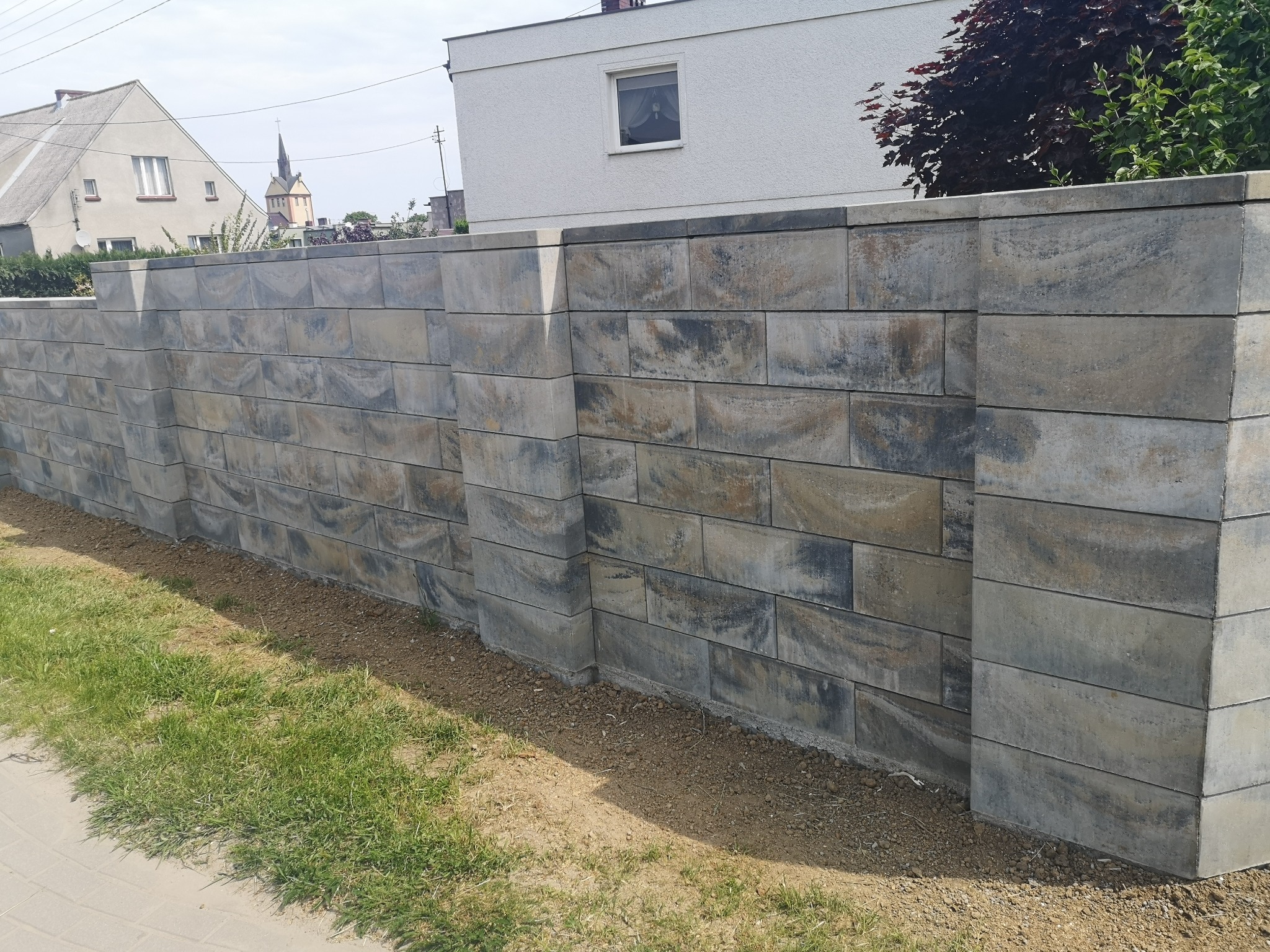 Nowoczesny mur z szarych, cieniowanych bloków betonowych, z widocznym tłem zabudowy jednorodzinnej i zielenią trawnika u podstawy.
