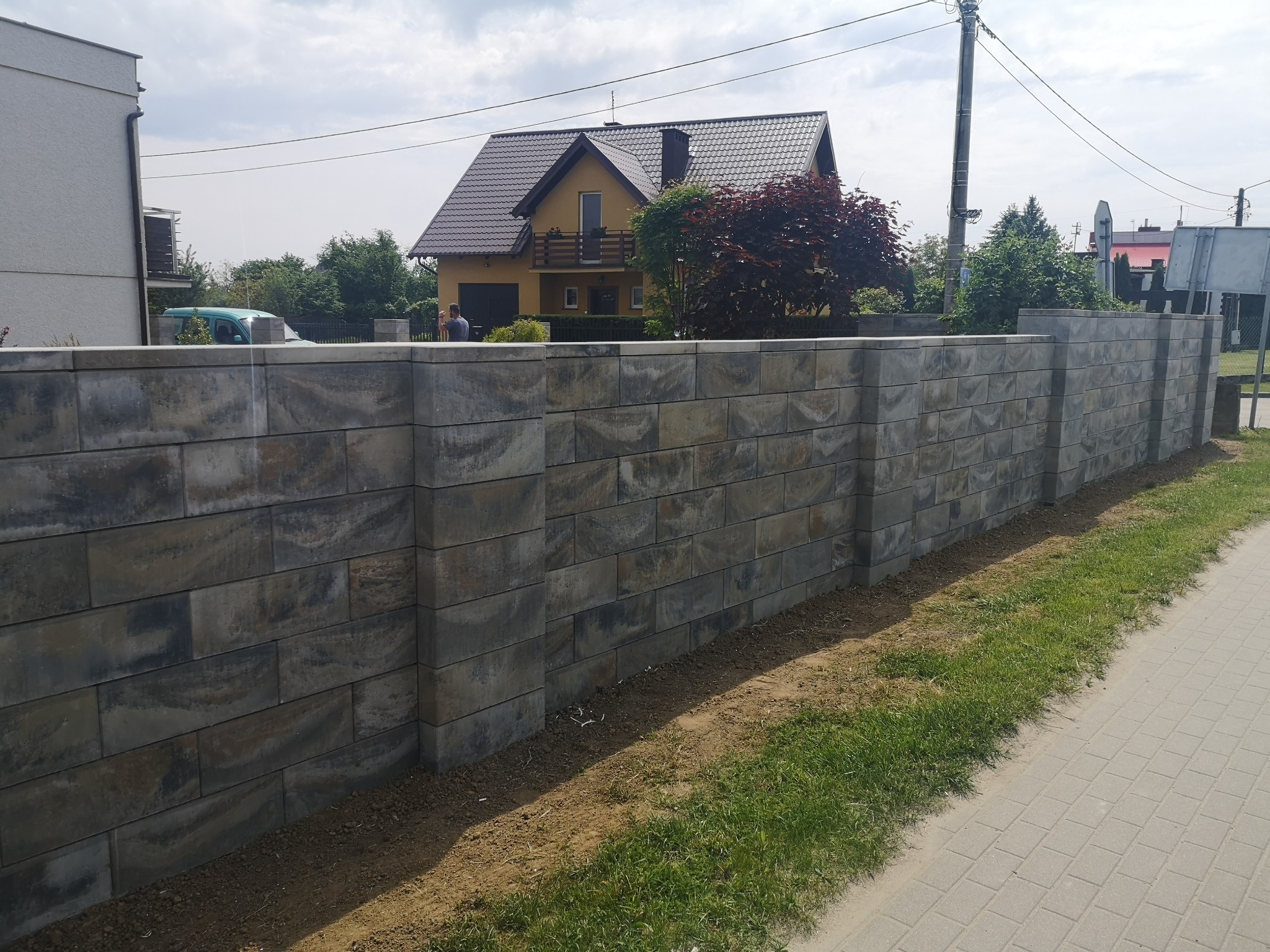 Nowoczesny, wysoki mur z szarej kostki łupanej, oddzielający posesję z żółtym domem z brązowym dachem, widoczny z chodnika wyłożonego kostką brukową.
