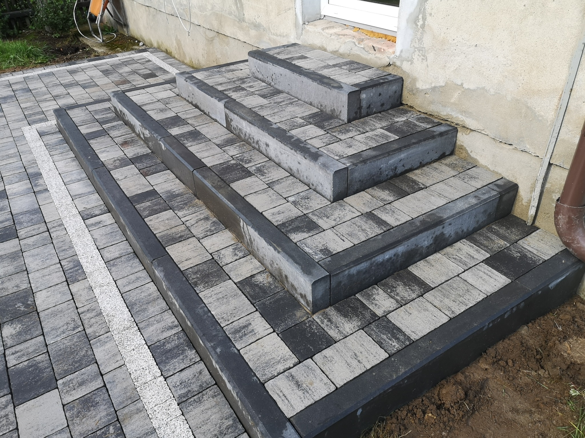 Schody zewnętrzne wyłożone szaro-czarną kostką brukową z ciemnoszarym obrzeżem, prowadzące do wejścia budynku, z jasnym pasem żwiru oddzielającym chodnik.