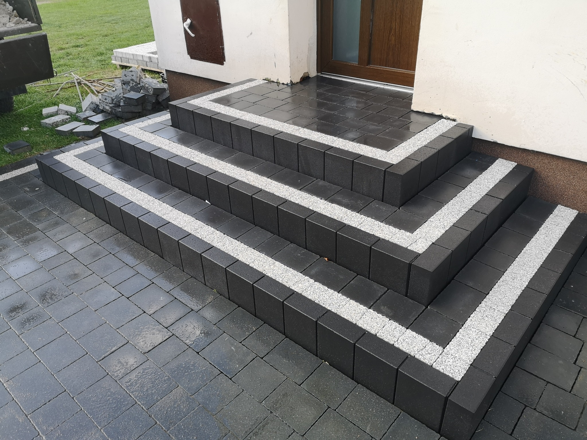 Schody wejściowe wyłożone ciemną kostką brukową z jasnymi akcentami, prowadzące do drzwi wejściowych domu.