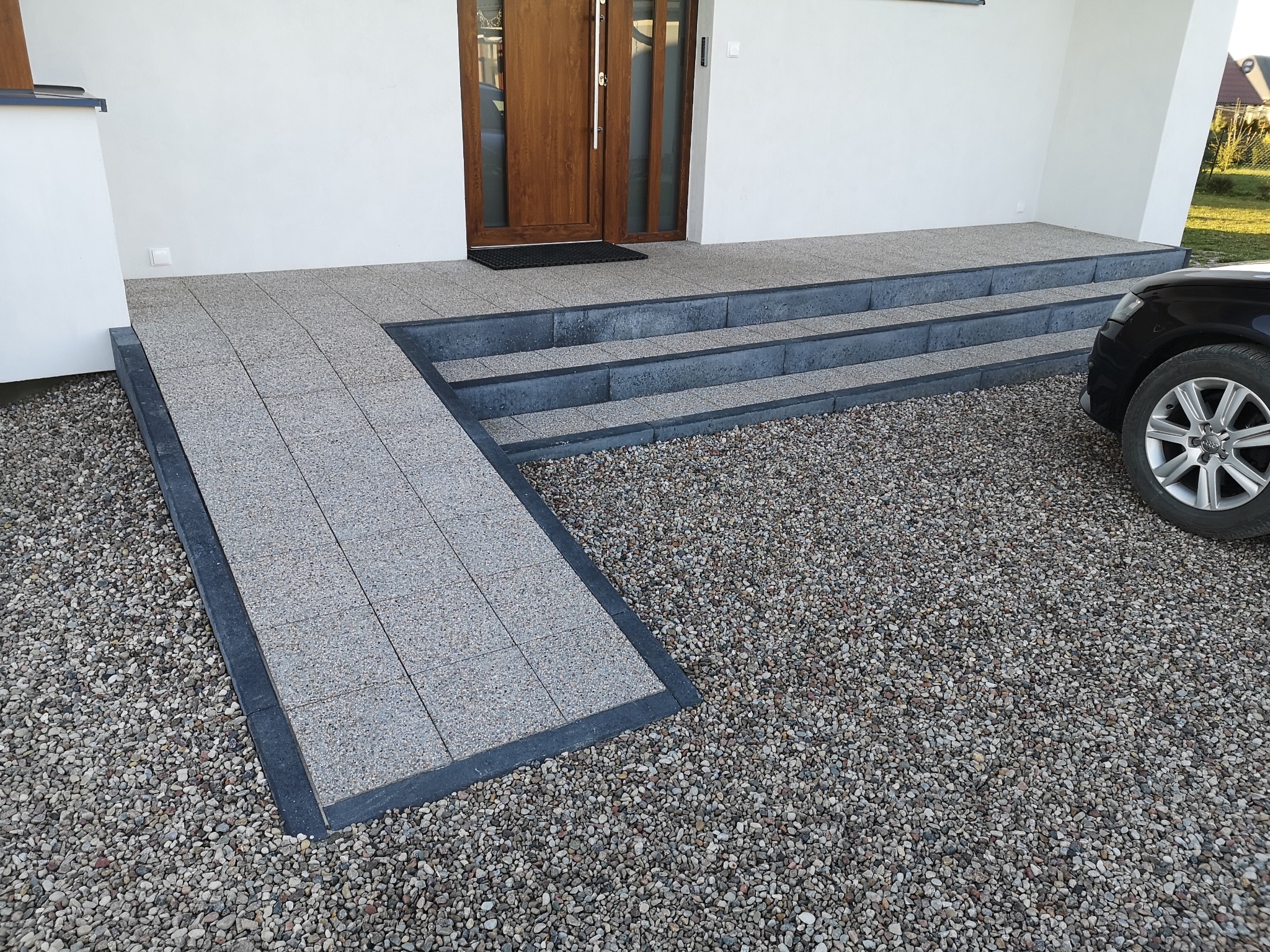 Nowoczesne wejście do domu z rampą i schodami wyłożonymi szarą kostką brukową, otoczone drobnym żwirem i fragmentem czarnego samochodu.