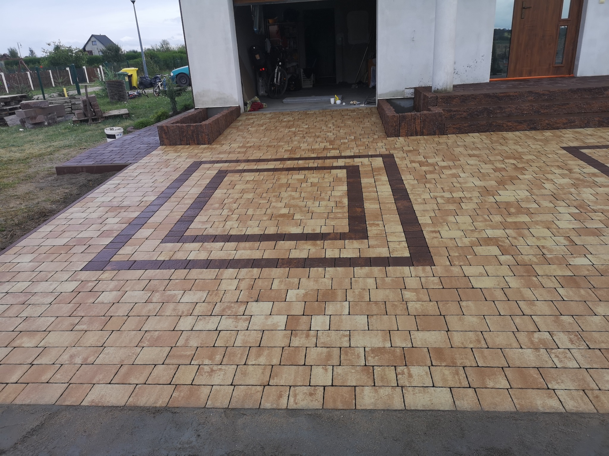 Ułożona kostka brukowa w geometryczny wzór przed domem z otwartym garażem, widoczne donice z kostki po bokach podjazdu i fragment ogrodu z tyłu.