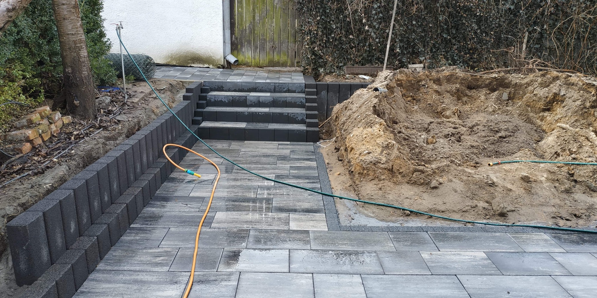 Ułożona szara kostka brukowa prowadząca do schodów z kostki, obok wykopana ziemia i ściana z bloczków betonowych, widoczne węże ogrodowe.