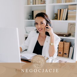 Karolina Czernecka-Cholewa - Uśmiechnięta kobieta w białej marynarce rozmawia przez telefon przy biurku z otwartym notatnikiem i komputerem; w tle regał z książkami.