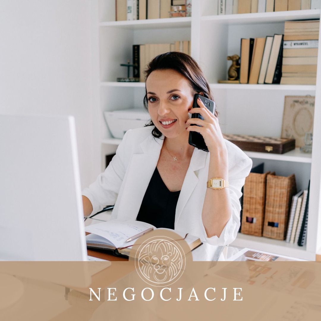 Uśmiechnięta kobieta w białej marynarce rozmawia przez telefon przy biurku z otwartym notatnikiem i komputerem; w tle regał z książkami.