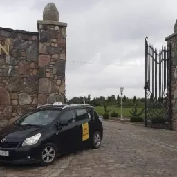 Czarny samochód taxi marki Toyota z żółtą naklejką na drzwiach, zaparkowany przed kamienną bramą z napisem KISTON i trzema gwiazdkami