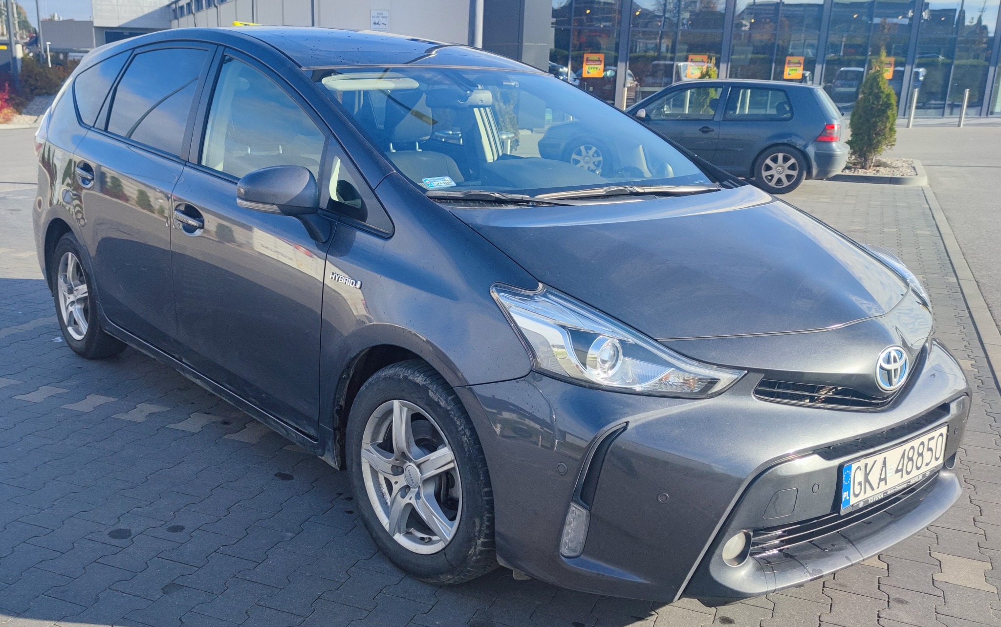 Szary minivan Toyota Prius Plus zaparkowany na kostce brukowej przed budynkiem z dużymi oknami, widoczna tablica rejestracyjna GKA 48850.