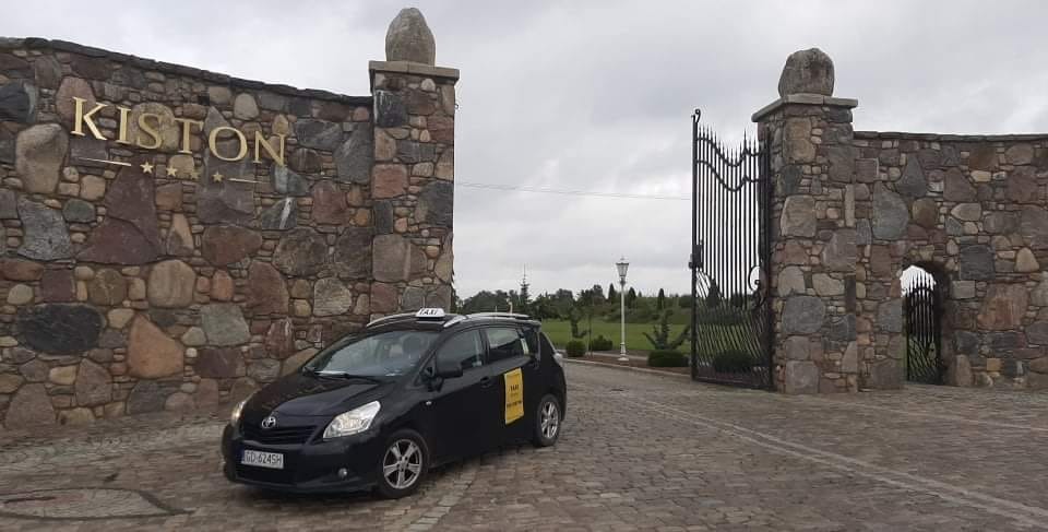 Czarny samochód taxi marki Toyota z żółtą naklejką na drzwiach, zaparkowany przed kamienną bramą z napisem KISTON i trzema gwiazdkami