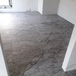 Szare płytki podłogowe imitujące marmur w surowym wnętrzu, widoczny przew&oacute;d elektryczny wystający z podłogi, okno z PCV w tle.