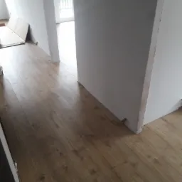 Ułożone panele podłogowe w jasnym odcieniu drewna, widoczne niedokończone ściany i kartony w tle, fragment balkonu w oddali.