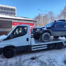 Laweta przewozi terenowy samochód Jeep Grand Cherokee z dodatkowym kołem zapasowym na dachu, na tle budynku warsztatu Motrio i błękitnego nieba.
