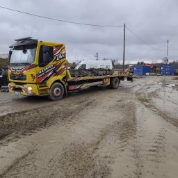 Transport Małgorzata Kraczkowska - Żółta laweta z napisem 'Transfer Pomoc Drogowa 24h' na błotnistym terenie. W tle kontenery i inne pojazdy. Pochmurne niebo.