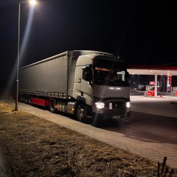 Transport Małgorzata Kraczkowska - Ciężarówka z naczepą na parkingu stacji benzynowej nocą, oświetlona latarnią. Widoczny numer rejestracyjny i dystrybutory paliw. Ciemne tło.