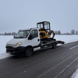 Transport Małgorzata Kraczkowska - Laweta z żółtą mini koparką na platformie, zaparkowana na poboczu ośnieżonej drogi. Widoczny biały samochód Iveco z numerem rejestracyjnym. Zimowy krajobraz w tle.