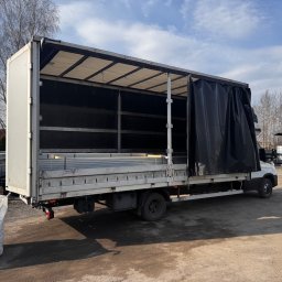 Transport Małgorzata Kraczkowska - Otwarty samochód dostawczy z plandeką, załadowany metalowymi profilami. Obok duży worek i budynek w tle. Widoczne elementy konstrukcyjne.
