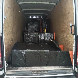 Wnętrze furgonetki wyłożone sklejką, wypełnione meblami zabezpieczonymi czarną folią, widoczne z tyłu pojazdu.