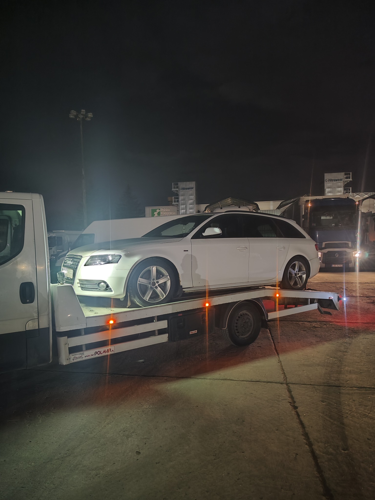 Białe kombi Audi A4 na lawecie w nocy, widoczny fragment kabiny lawety i oświetlenie uliczne w tle.