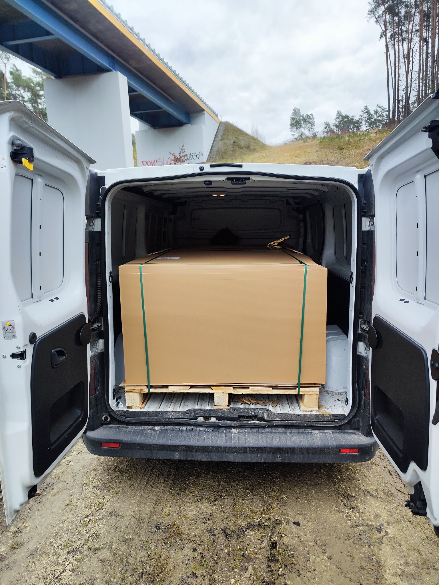 Wnętrze białego busa z otwartymi drzwiami, załadowane dużą kartonową paczką na palecie, zabezpieczoną zielonymi pasami; w tle widoczny most i drzewa.