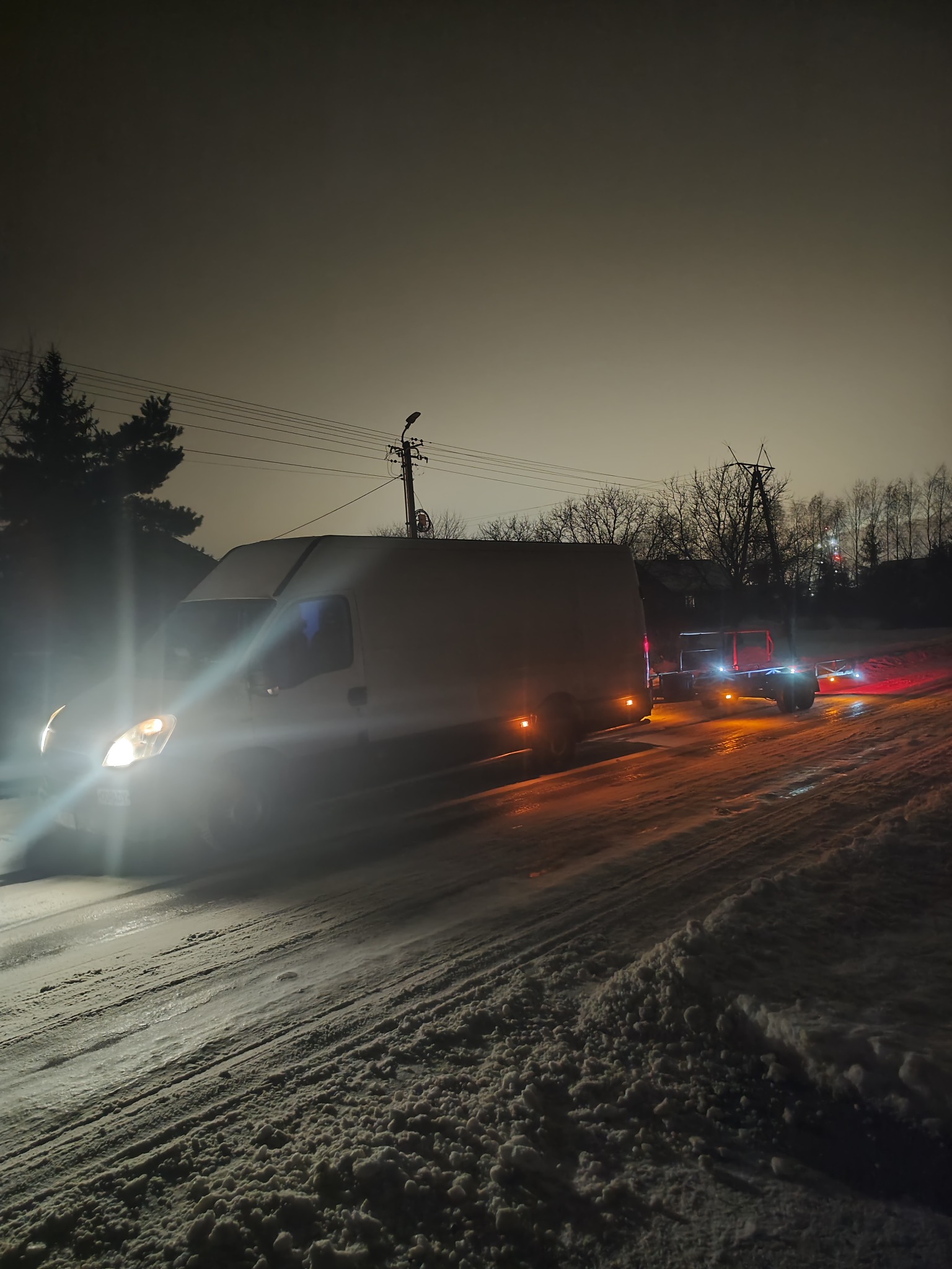 Biały van z przyczepą na ośnieżonej drodze w nocy, widoczne światła pojazdów i oświetlenie uliczne